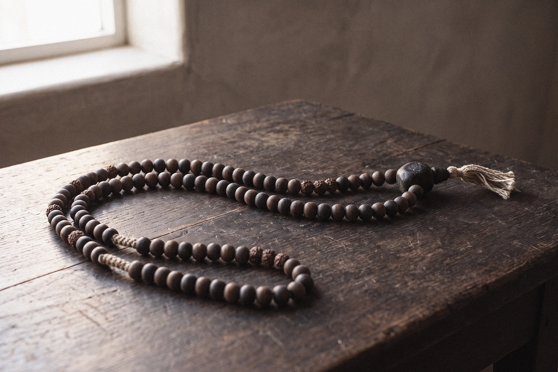 A long strand of dark mala beads laid out across a dark weathered wooden table, with a large guru bead and a pale tassel at one end.