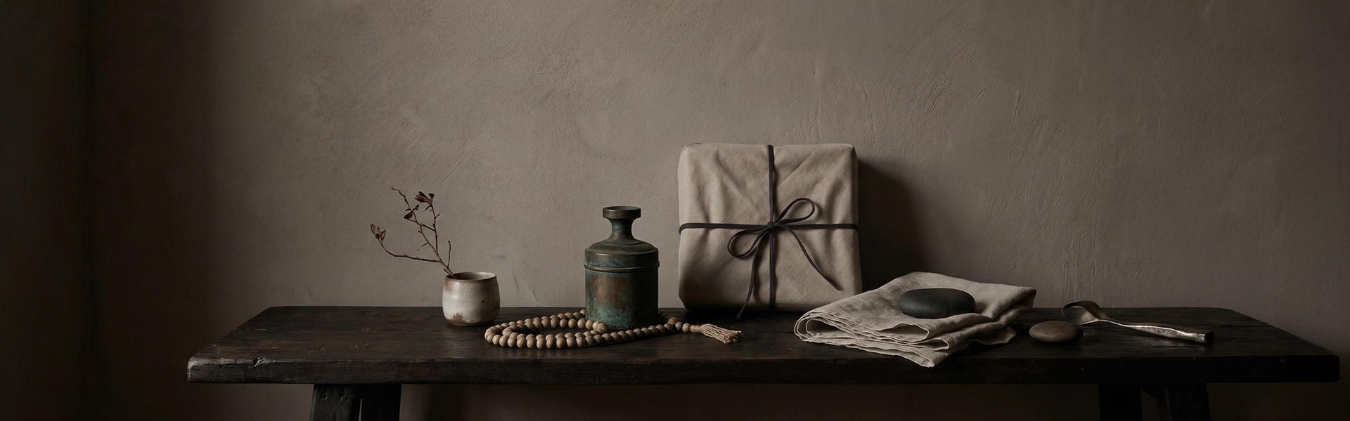 Gift ritual scene with a linen-wrapped package, muted bead strand, aged metal vessel, ceramic cup, folded cloth, raw stone, and quiet wabi-sabi gift atmosphere.