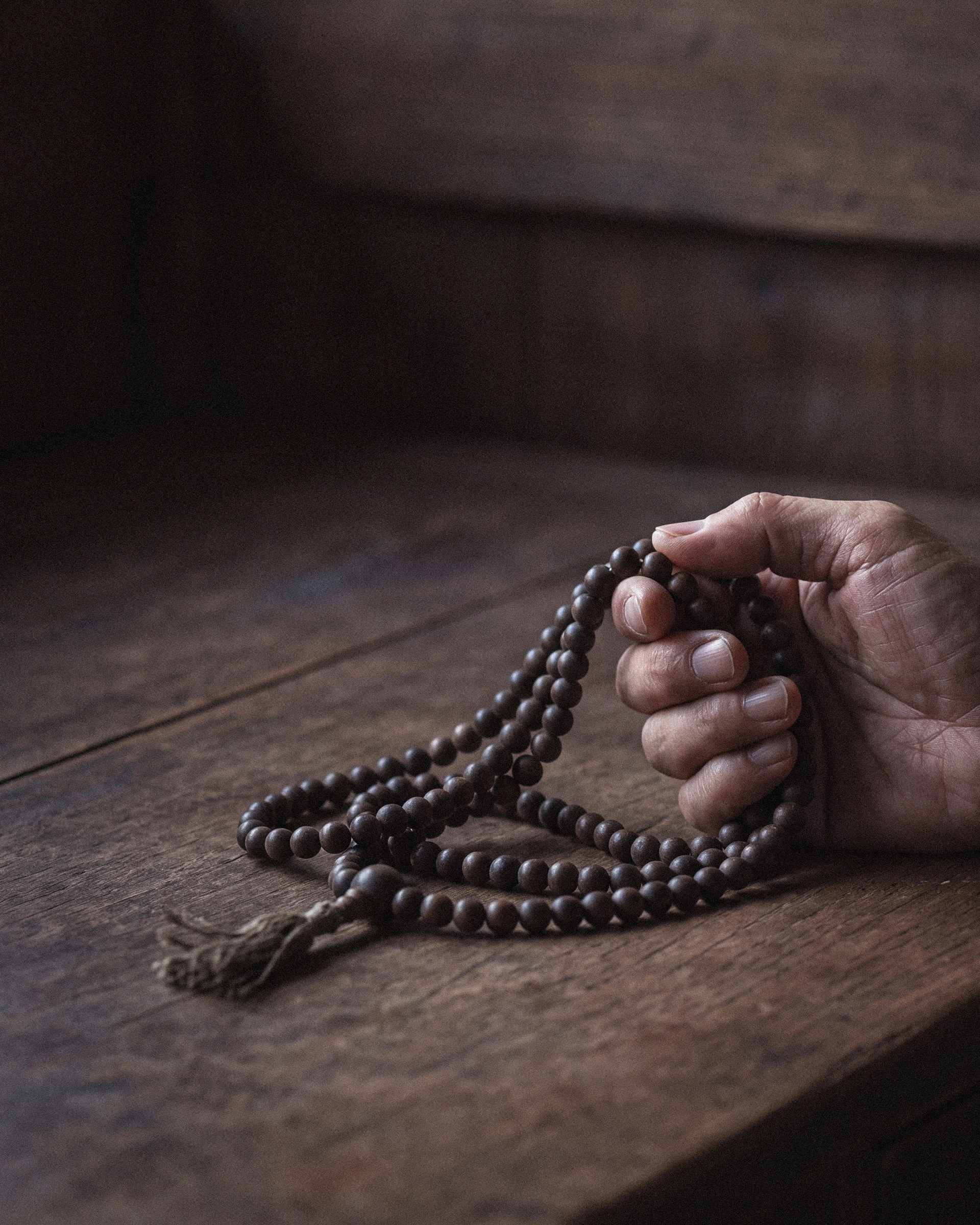 A hand holding a looped strand of dark mala beads above a wooden surface, with the tassel resting on the wood.