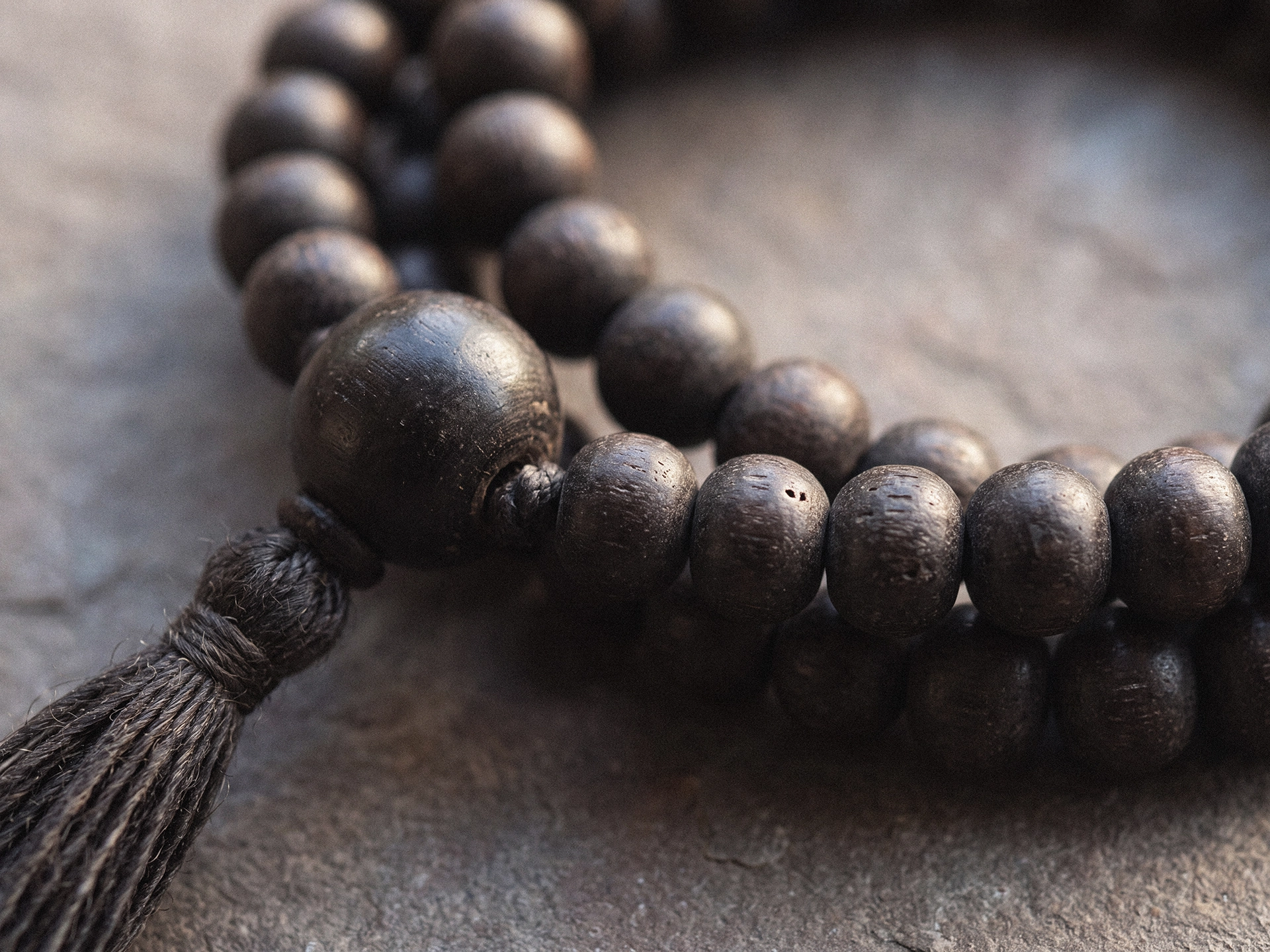 A close-up of the head of a dark-beaded mala, showing a large rounded guru bead, its knotted cord, and the beginning of a dark tassel.