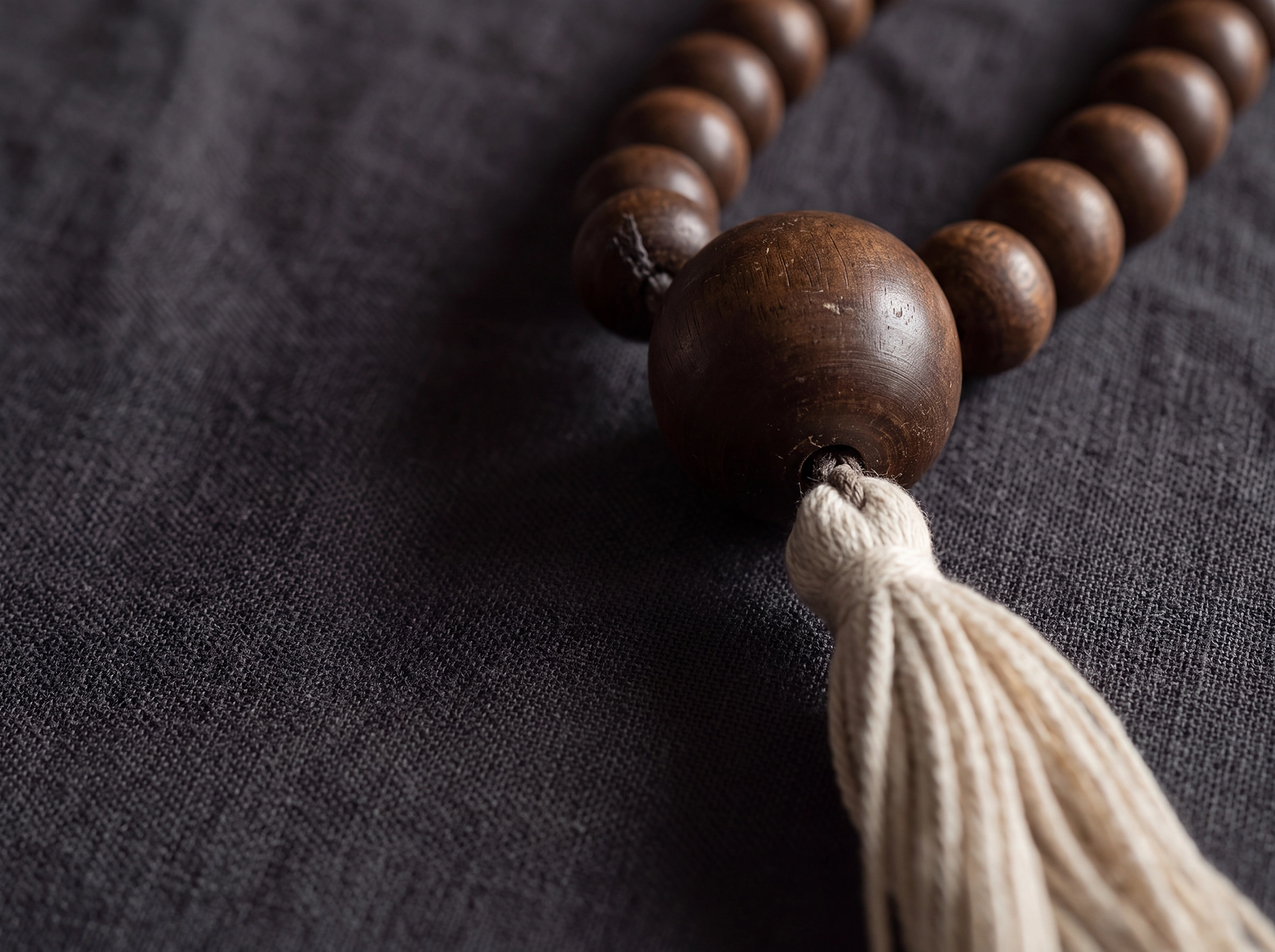 Close-up of a mala strand's junction, showing a larger dark wooden guru bead with an ivory cotton tassel hanging below it on dark linen.