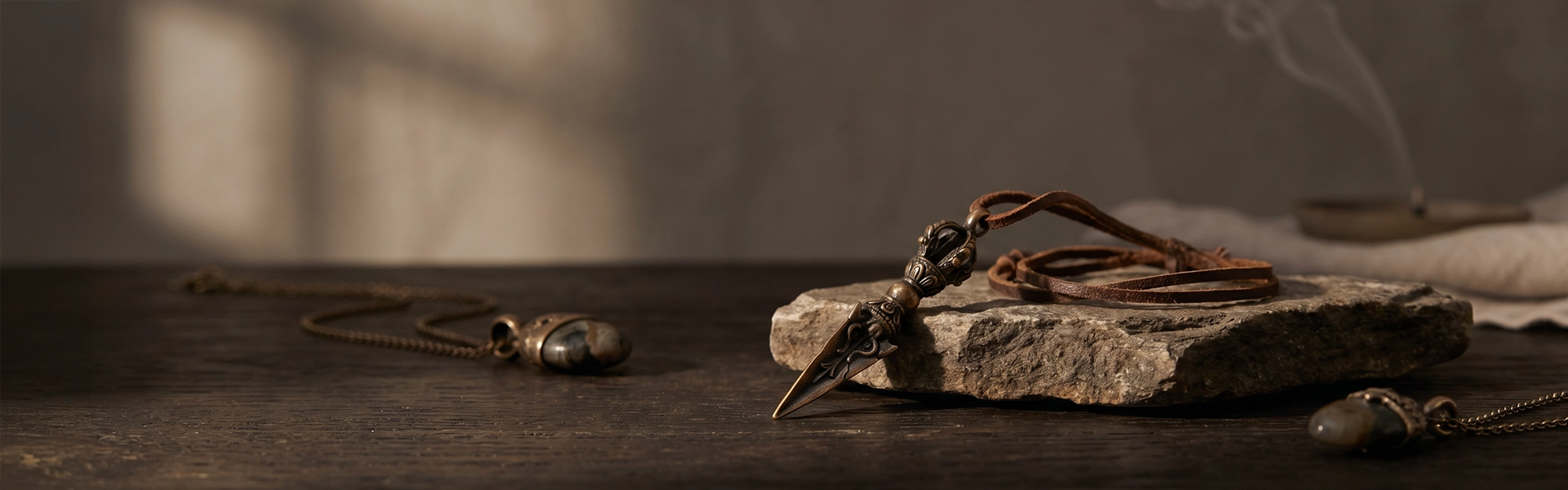 Phurba pendant necklace arranged on raw stone and dark wood in a quiet Western wabi-sabi ritual setting with aged metal, soft smoke, and warm shadow.