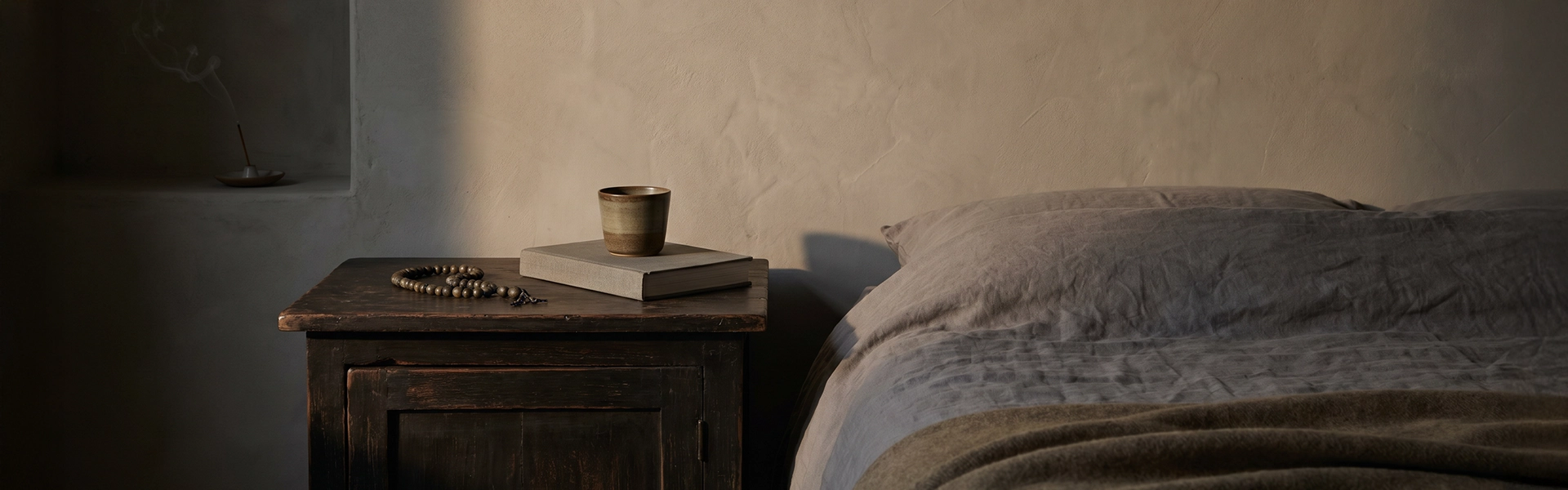 Nightstand and bedside scene with muted beads, ceramic cup, closed book, linen bedding, and quiet evening ritual atmosphere.