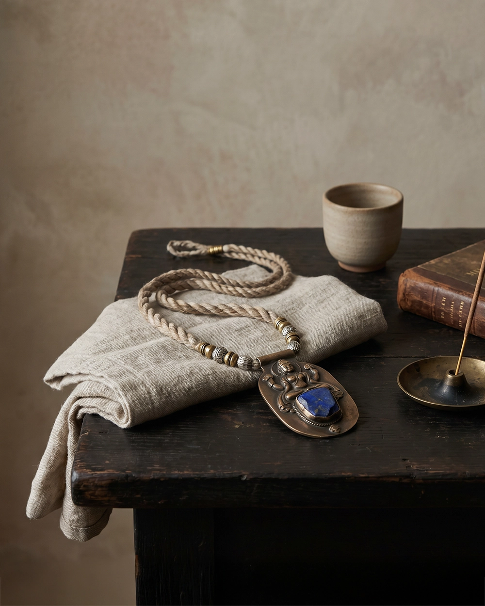 Himalayan-inspired pendant amulet on linen with incense, an old book, and a ceramic cup on a dark wooden table