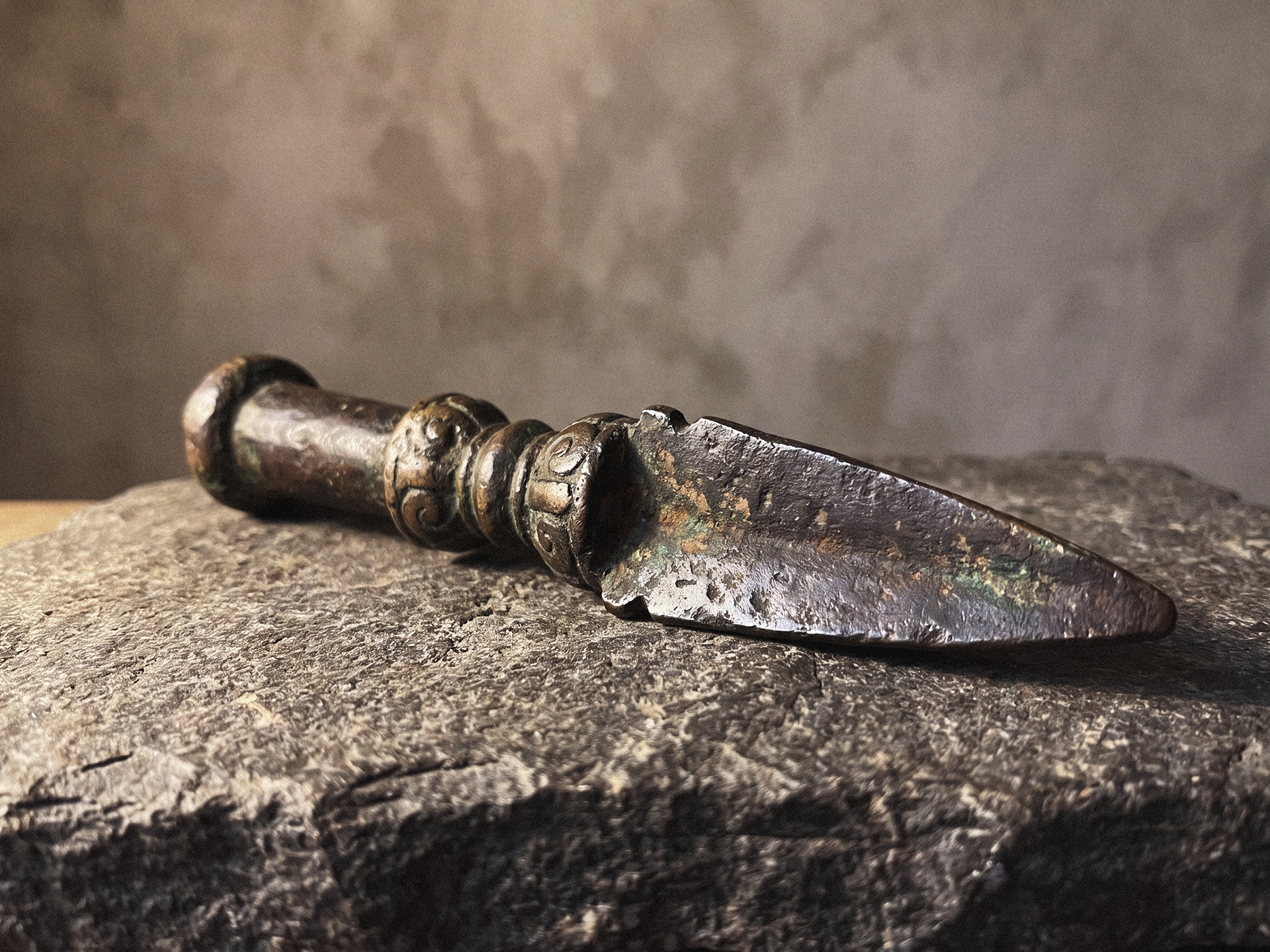 A close-up of a phurba-form object with a carved mid-section and tapering blade resting on a rough stone surface.