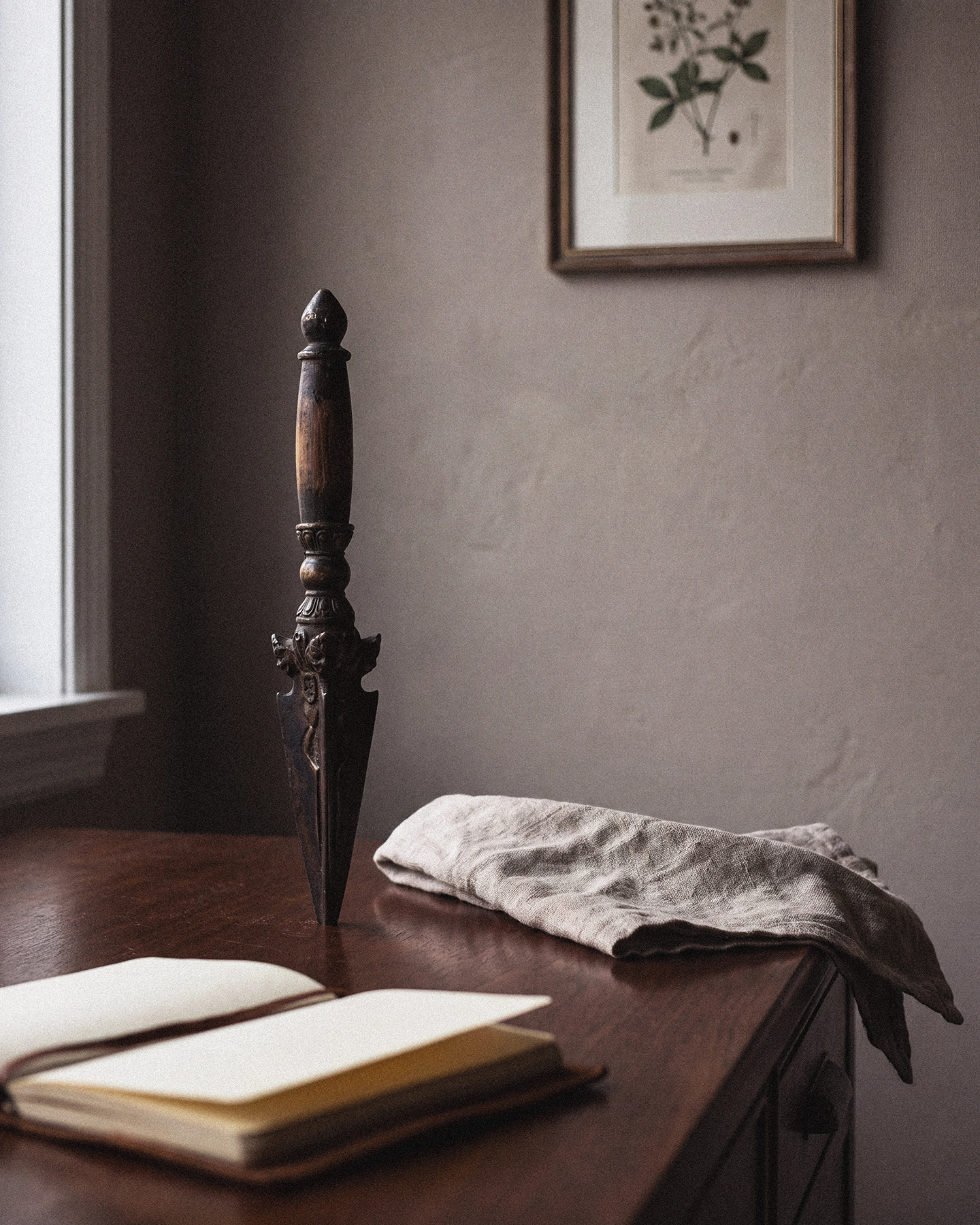 A phurba-form object standing upright on a dark wooden study desk beside a folded linen cloth and an open notebook.