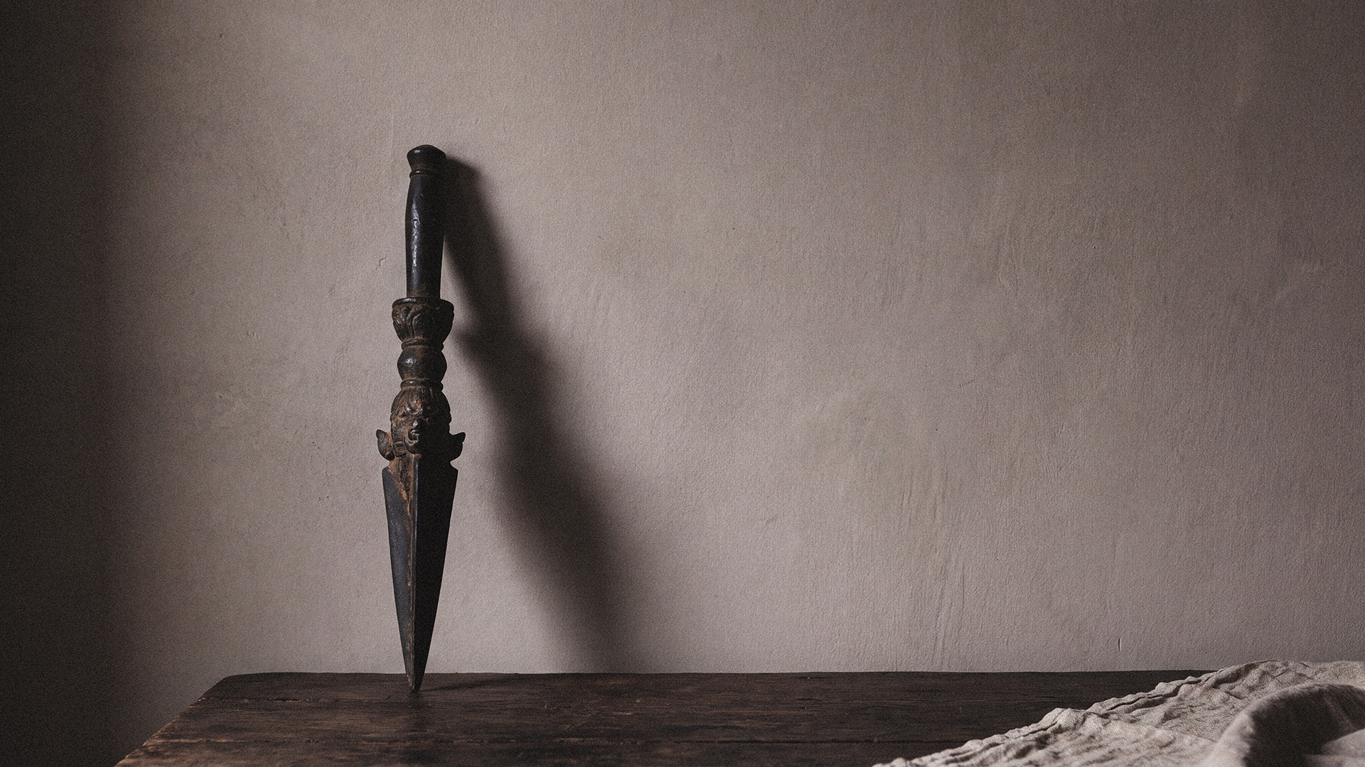 A dark phurba-form object with a carved handle standing upright on a dark wooden surface, casting a long shadow against a warm gray wall.