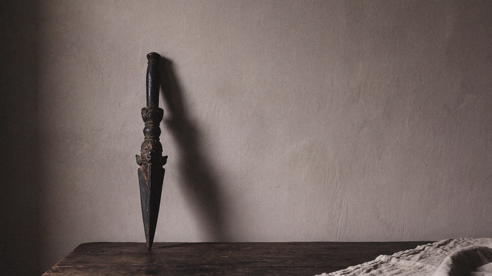A dark phurba-form object with a carved handle standing upright on a dark wooden surface, casting a long shadow against a warm gray wall.