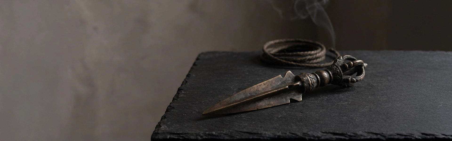 Phurba Kila inspired bronze ritual object arranged on black slate with dark cord, soft smoke, and quiet wabi-sabi atmosphere.
