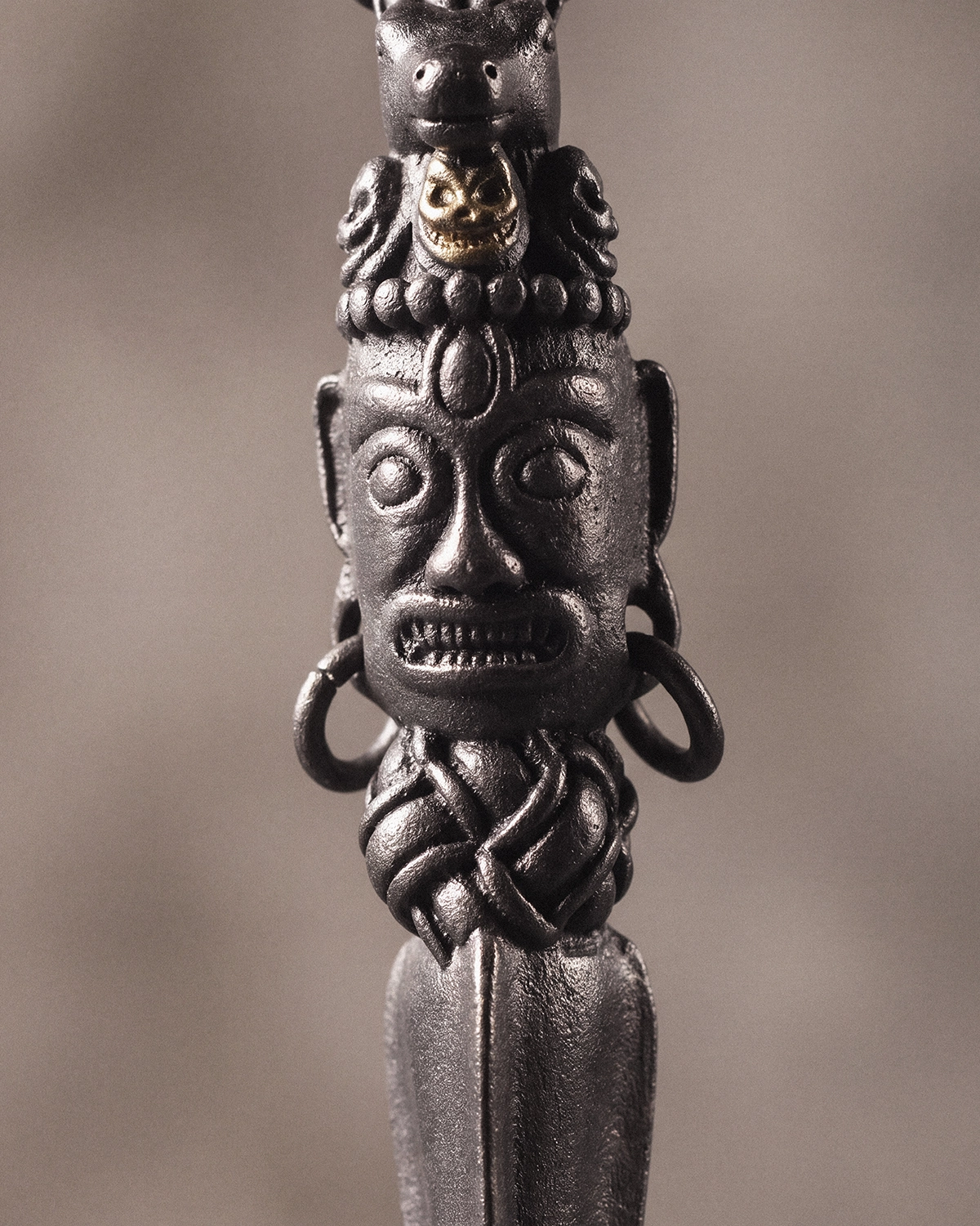 Close-up of a Phurba pendant showing a carved fierce guardian face, horse-head crown, small gold-toned skull detail, and dark oxidized metal texture.