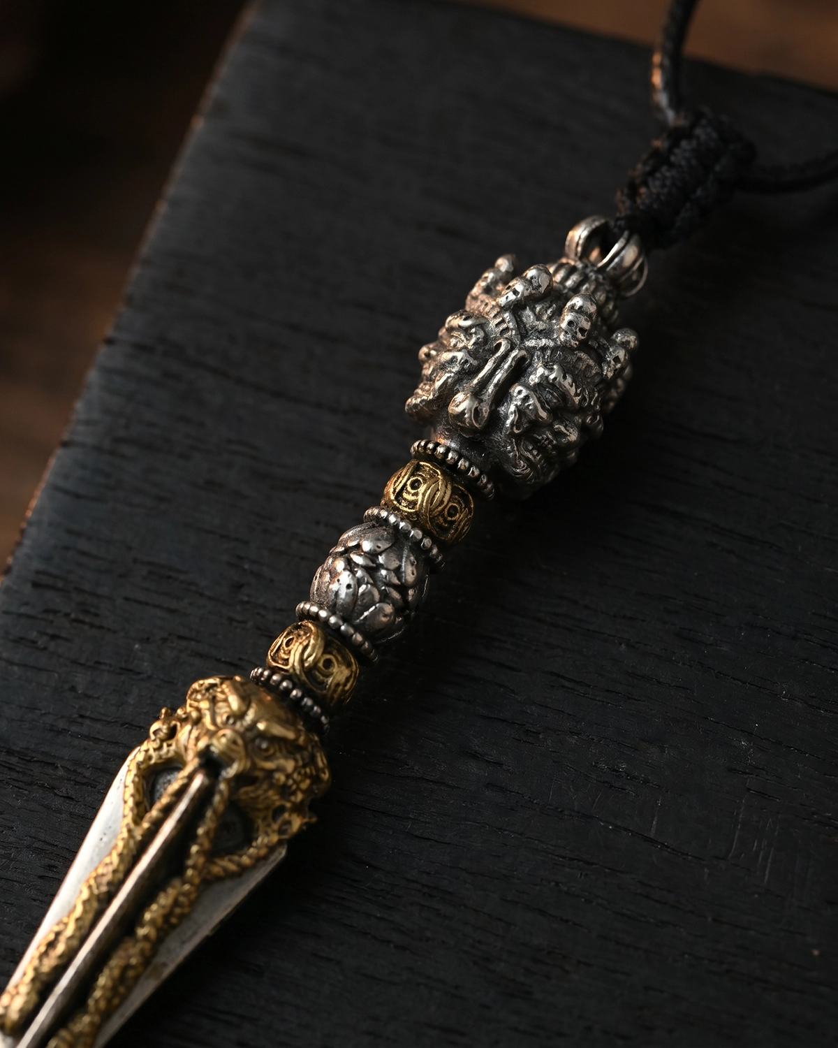 Close-up of a brass Phurba pendant necklace showing carved metal details and mixed silver- and gold-toned surfaces on a dark wood surface.