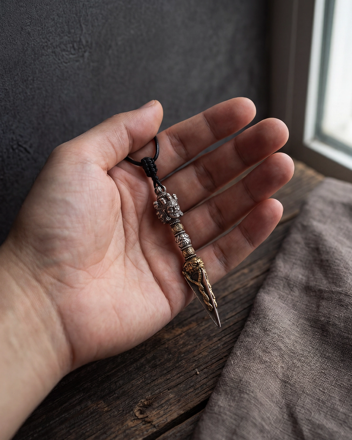 Brass Phurba pendant necklace held in an adult hand by a window to show its compact handheld scale.