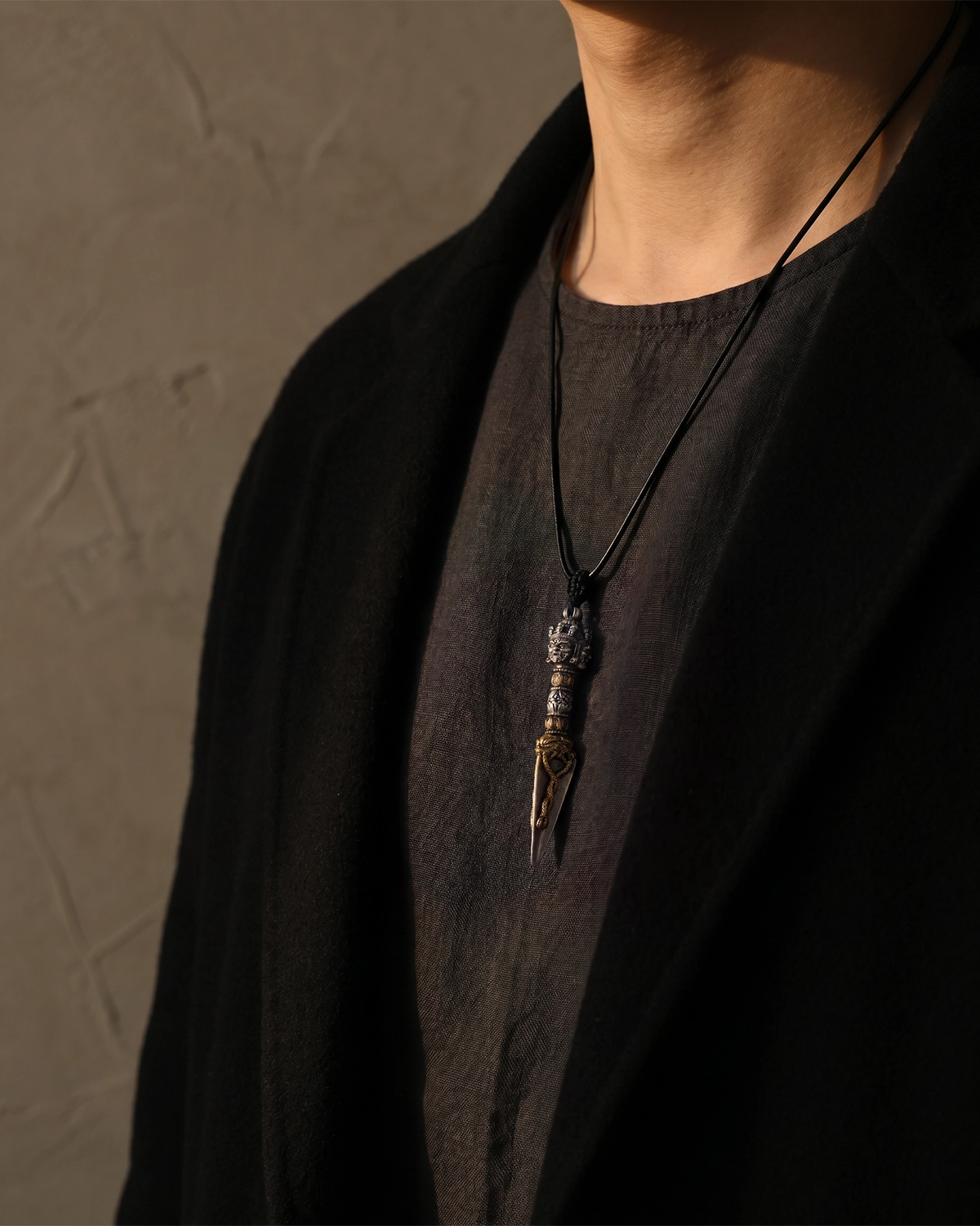 Brass Phurba pendant necklace worn on the upper chest with a black cord against a dark linen top and black outer layer.