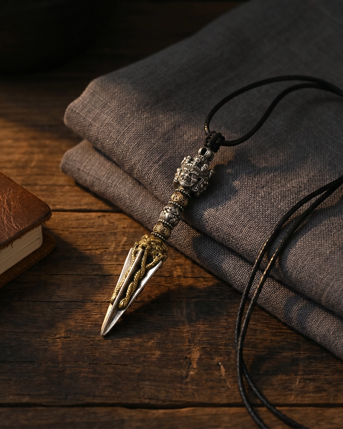 Brass Phurba pendant necklace resting on a wooden tabletop beside folded gray linen in a quiet still-life scene.