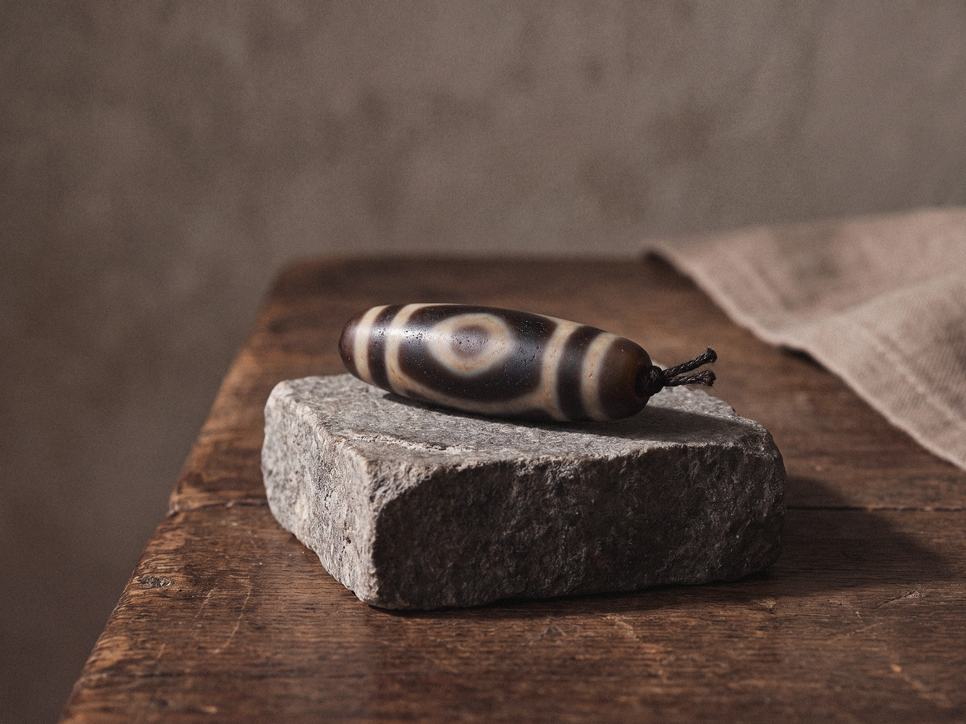 A single elongated dzi bead with banded eye-like markings resting on a rough gray stone block over aged wood.