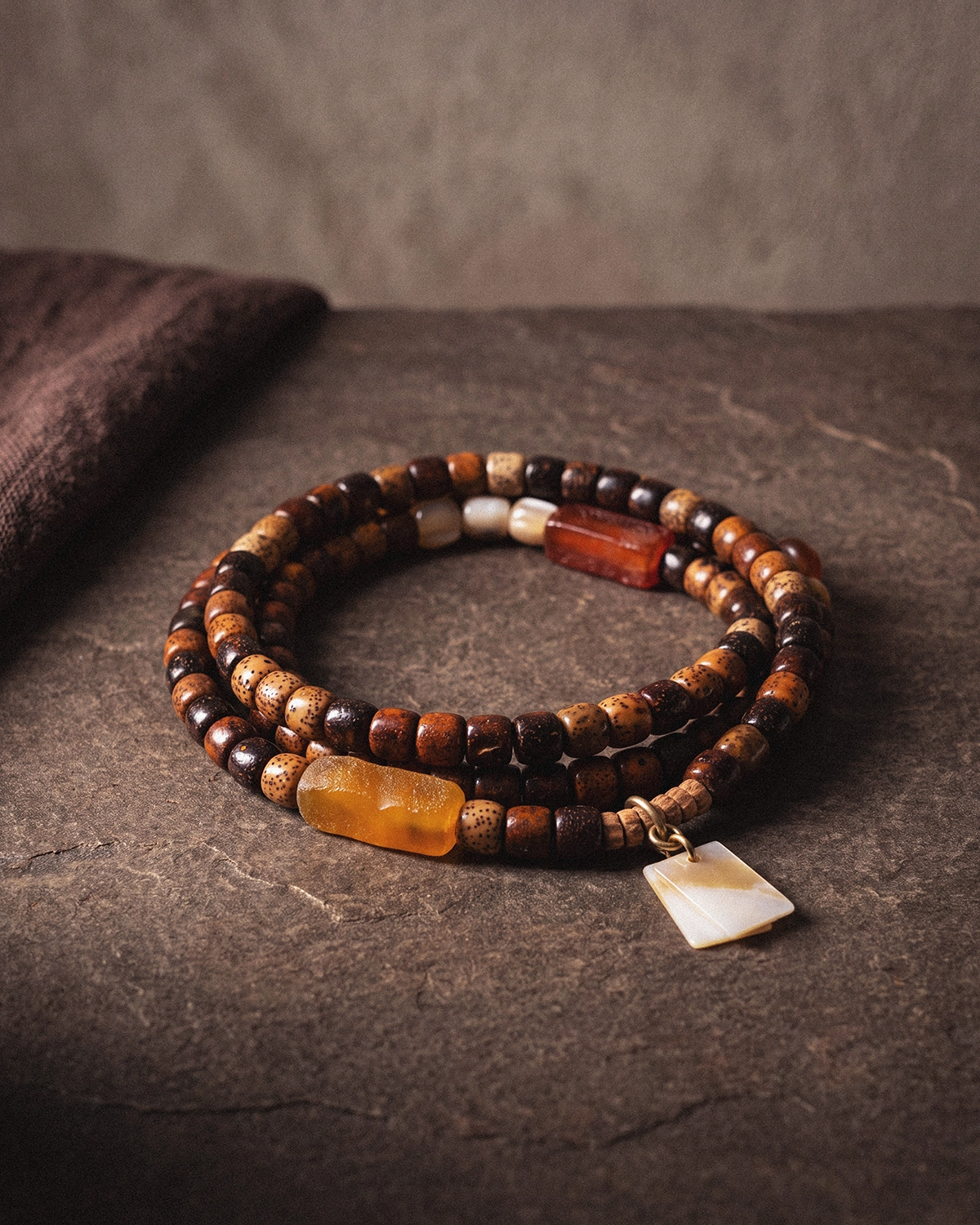 A 108-bead star and moon bodhi seed mala coiled in two loops on a slate-gray stone surface beside folded brown linen, with a shell pendant at the side.