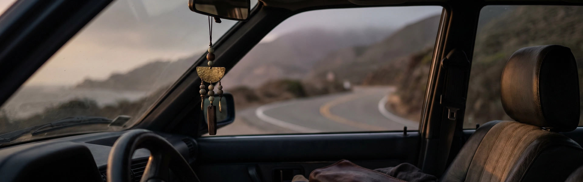 Travel and car scene with a rearview mirror charm, muted beads, aged metal, dark car interior, leather pouch, linen cloth, and a quiet road beyond the windshield.