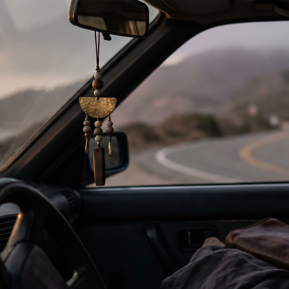 {"default":"Square mobile banner showing a rearview mirror charm with muted beads and aged metal inside a dark car interior, with a soft road landscape beyond the windshield."}
