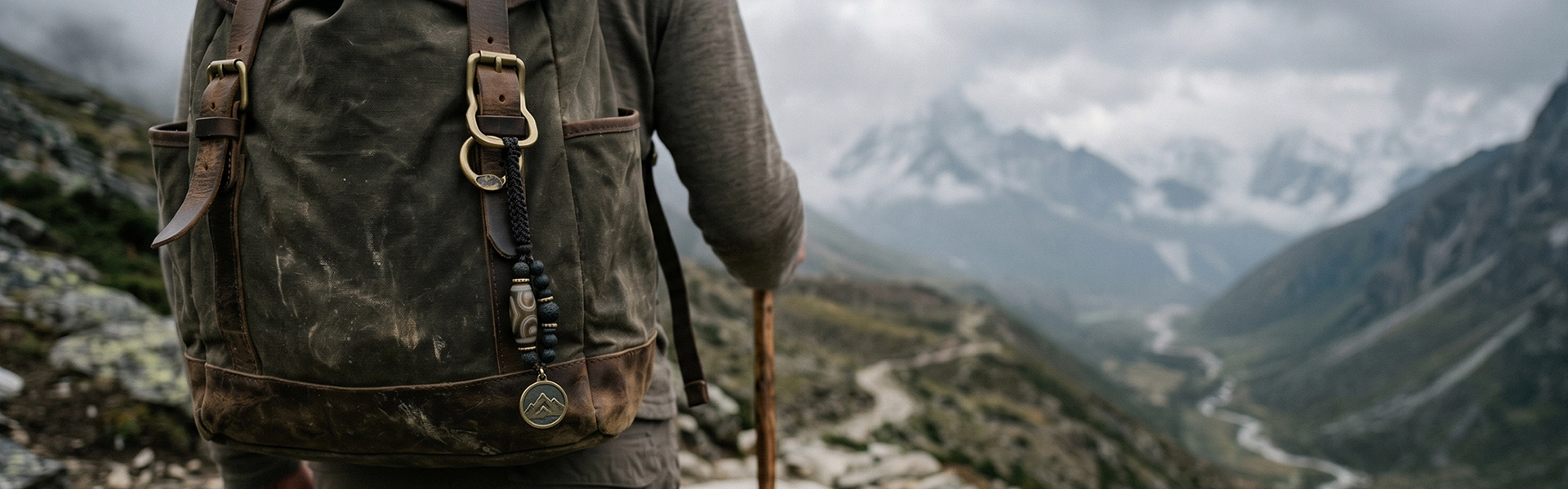 Tibetan-inspired travel charm with Dzi bead, dark cord, and aged metal pendant hanging from a hiking backpack on a misty mountain trail.