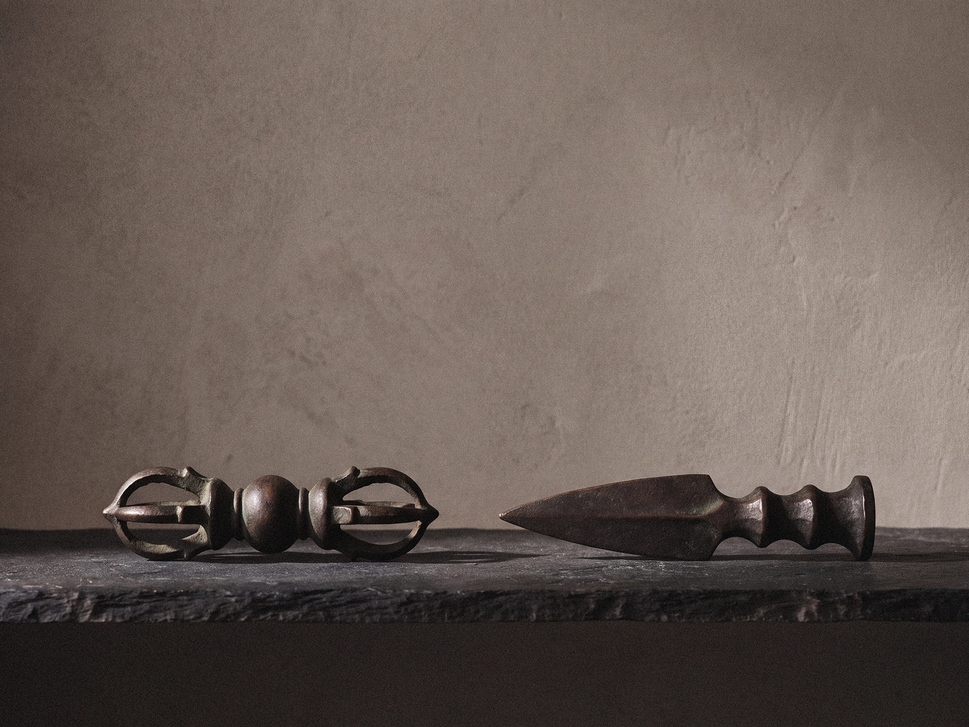 A short symmetrical double-ended vajra and a tapered phurba-form blade placed side by side on a dark stone ledge.