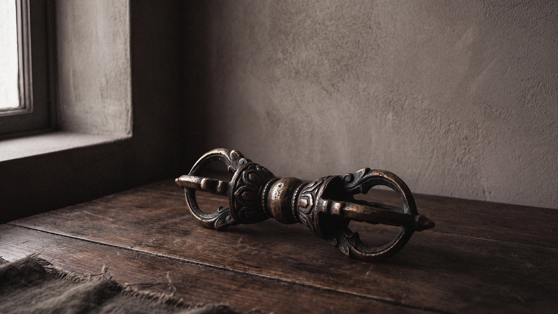 An aged-bronze vajra, also called a dorje, resting on a dark wooden surface beside a window in soft side light.