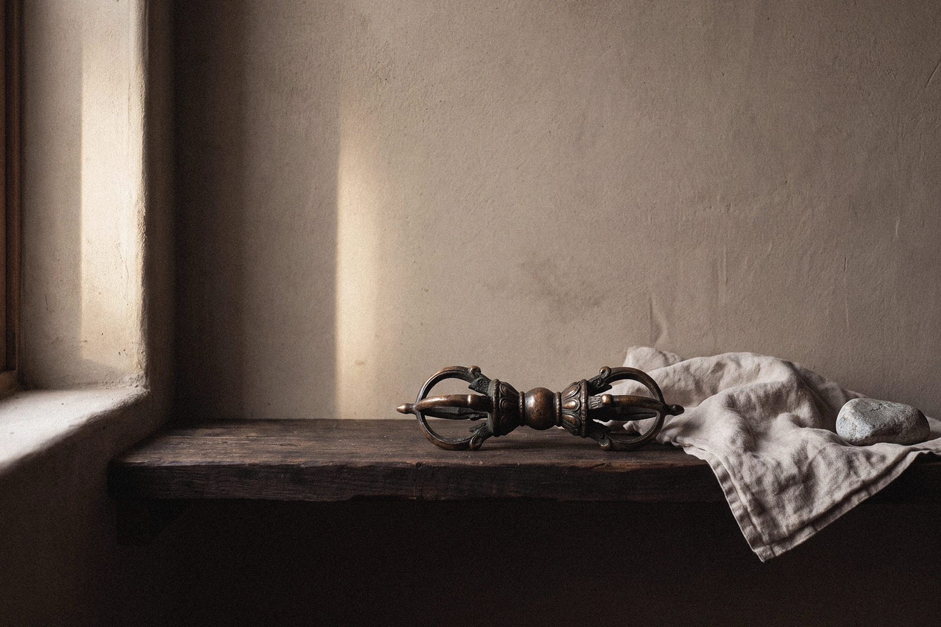 An aged-bronze vajra on a dark wood shelf beside draped linen cloth and a smooth stone under soft window light.