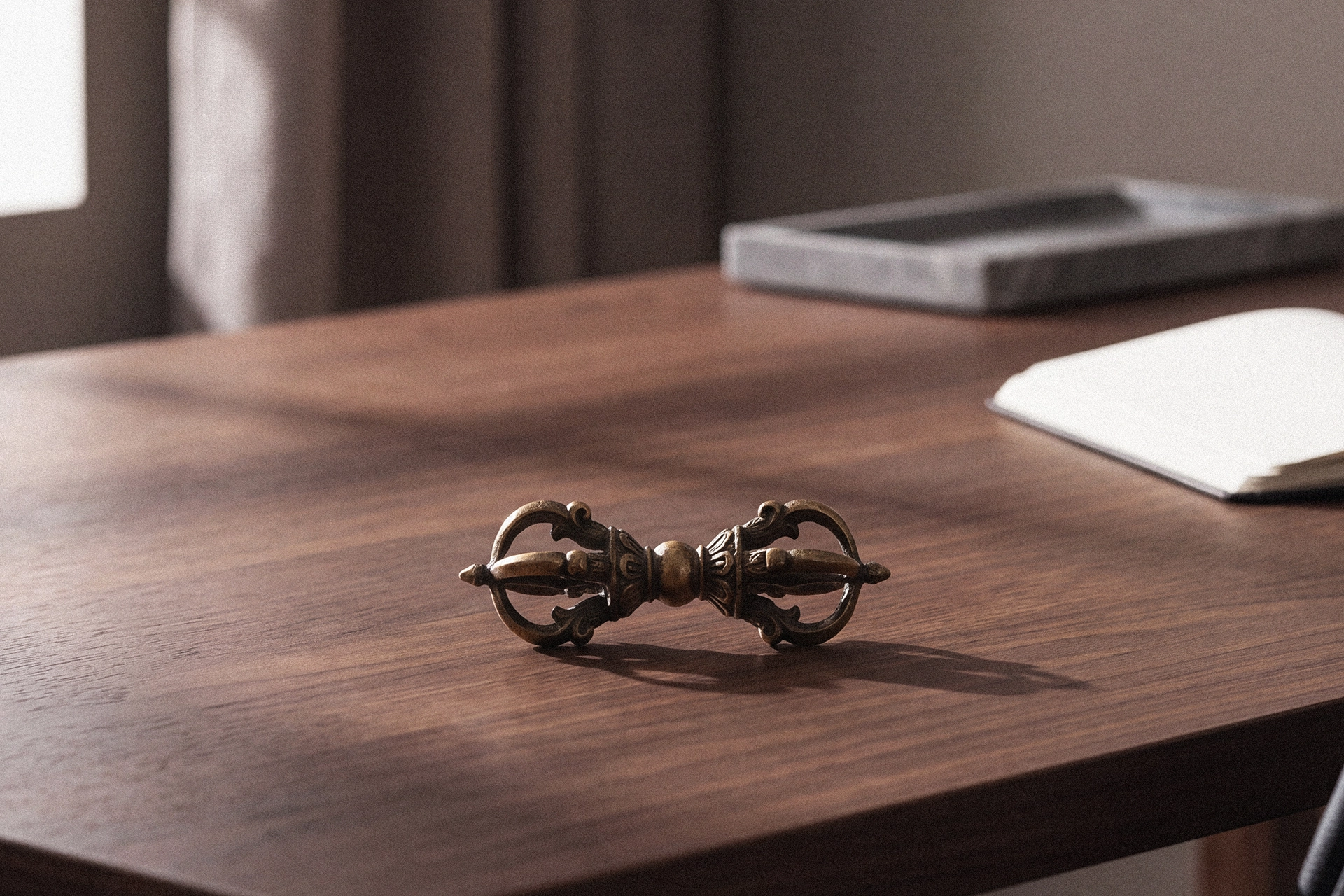 A small metal vajra on a dark wood desk, with a stone tray and an open notebook softly out of focus behind it.