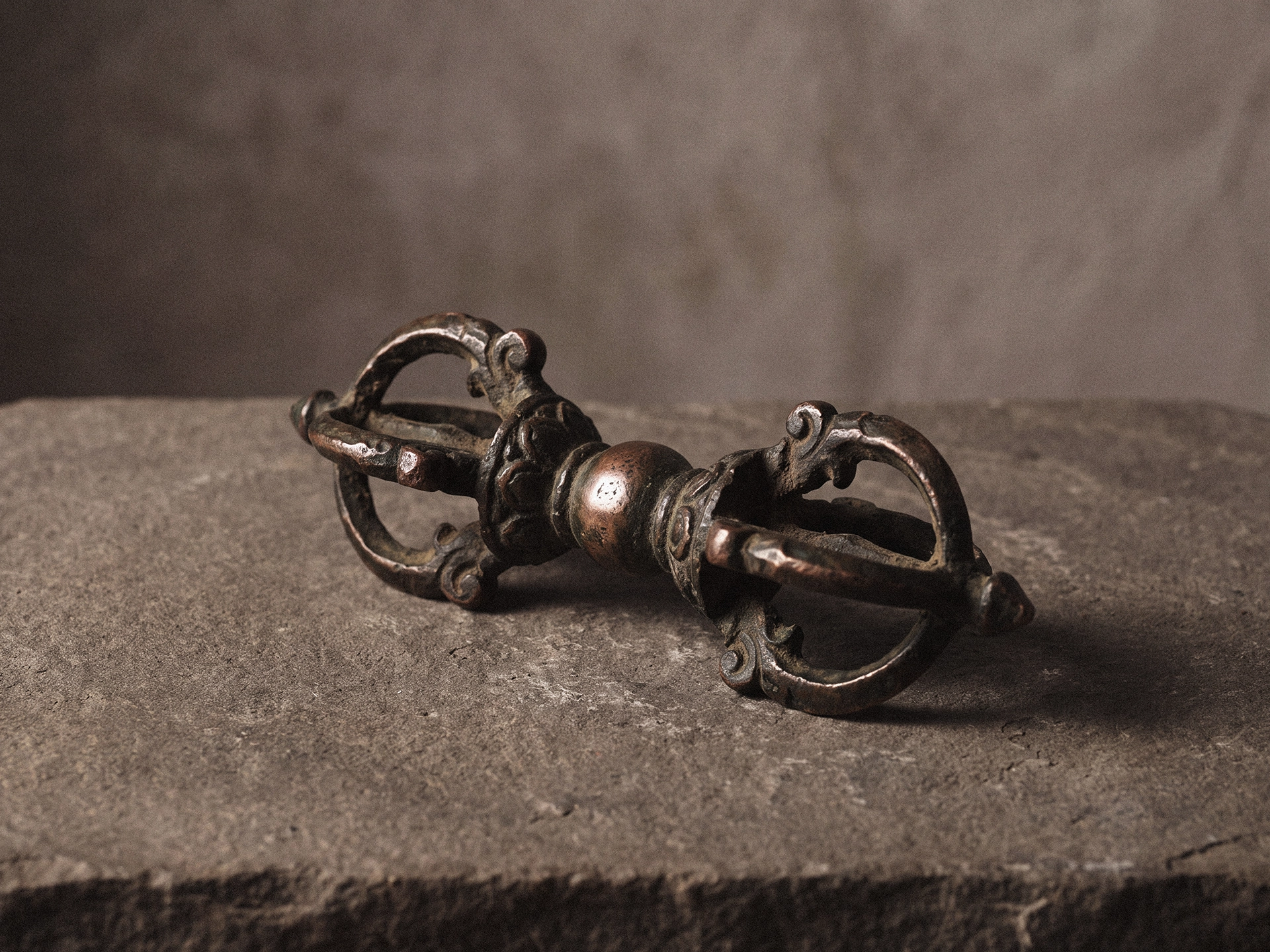 A double-ended vajra with a carved central grip and symmetrical prongs lying on a rough gray stone slab.