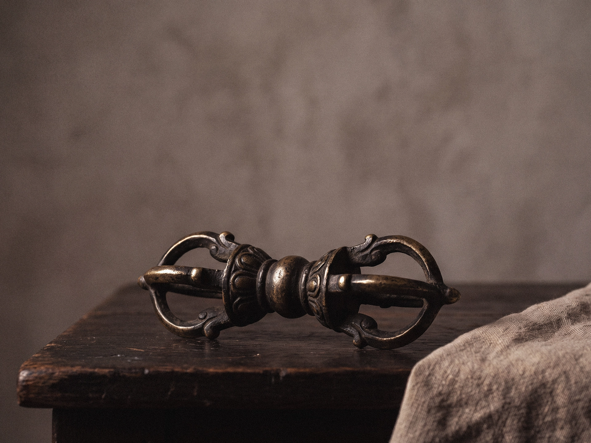 A close view of a metal vajra showing its central grip and symmetrical ornamental ends on dark wood, with linen at one edge.