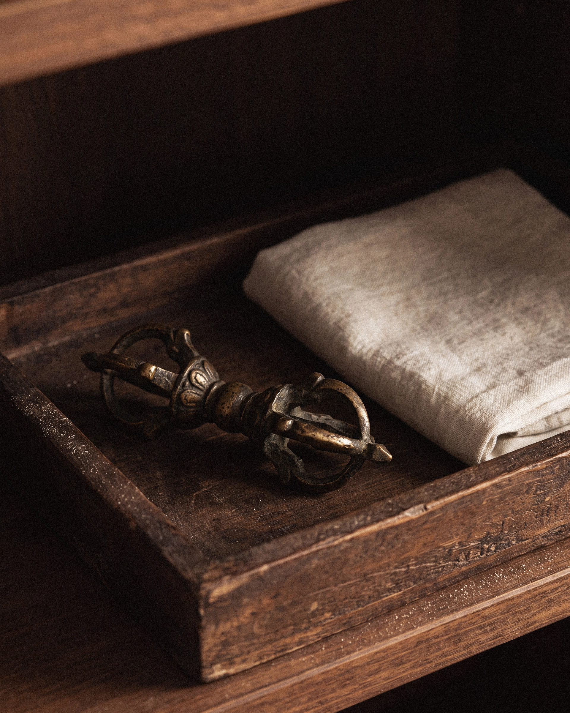 A metal vajra lying in a shallow dark wooden tray beside a neatly folded off-white linen cloth.