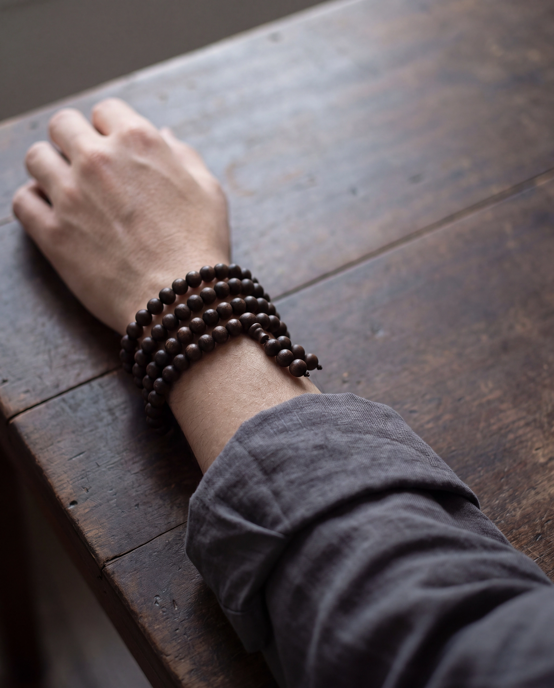 A dark wooden wrist mala wrapped several times around a wrist, with the arm resting on a dark wooden desk.