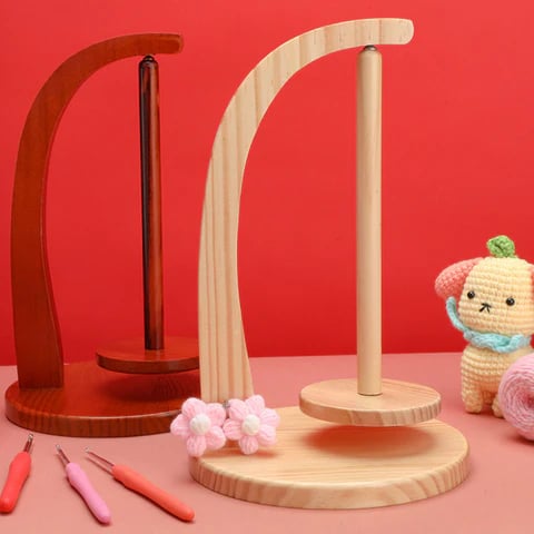The Levitating Premium Yarn Holder, featured on a red background with crochet hooks, pink yarn, and a crocheted bunny doll, offers seamless yarn management with its central spindle design.