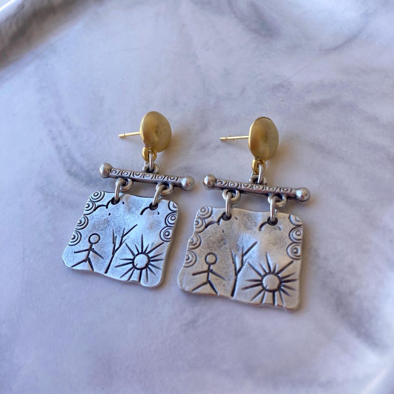 Close-up of a pair of handmade dangle earrings resting on a white marble surface. The earrings feature a mixed-metal design with yellow gold oval stud posts, a decorative silver horizontal bar, and a dangling square silver pendant engraved with ancient petroglyph-style symbols, including stick figures, trees, spirals, and a sun. The textured silver contrasts with the smooth gold. The earrings are photographed at a slightly angled top-down perspective in soft natural lighting.