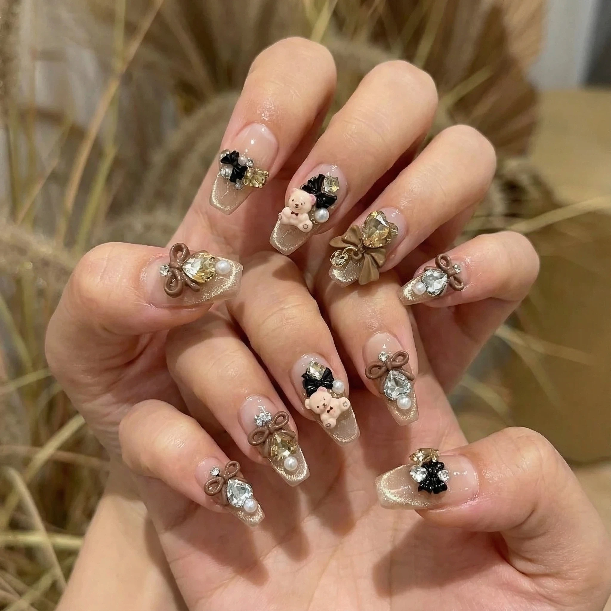 Hands wearing nude and black press-on nails with bow, pearl and rhinestone accents for a coquette-inspired look