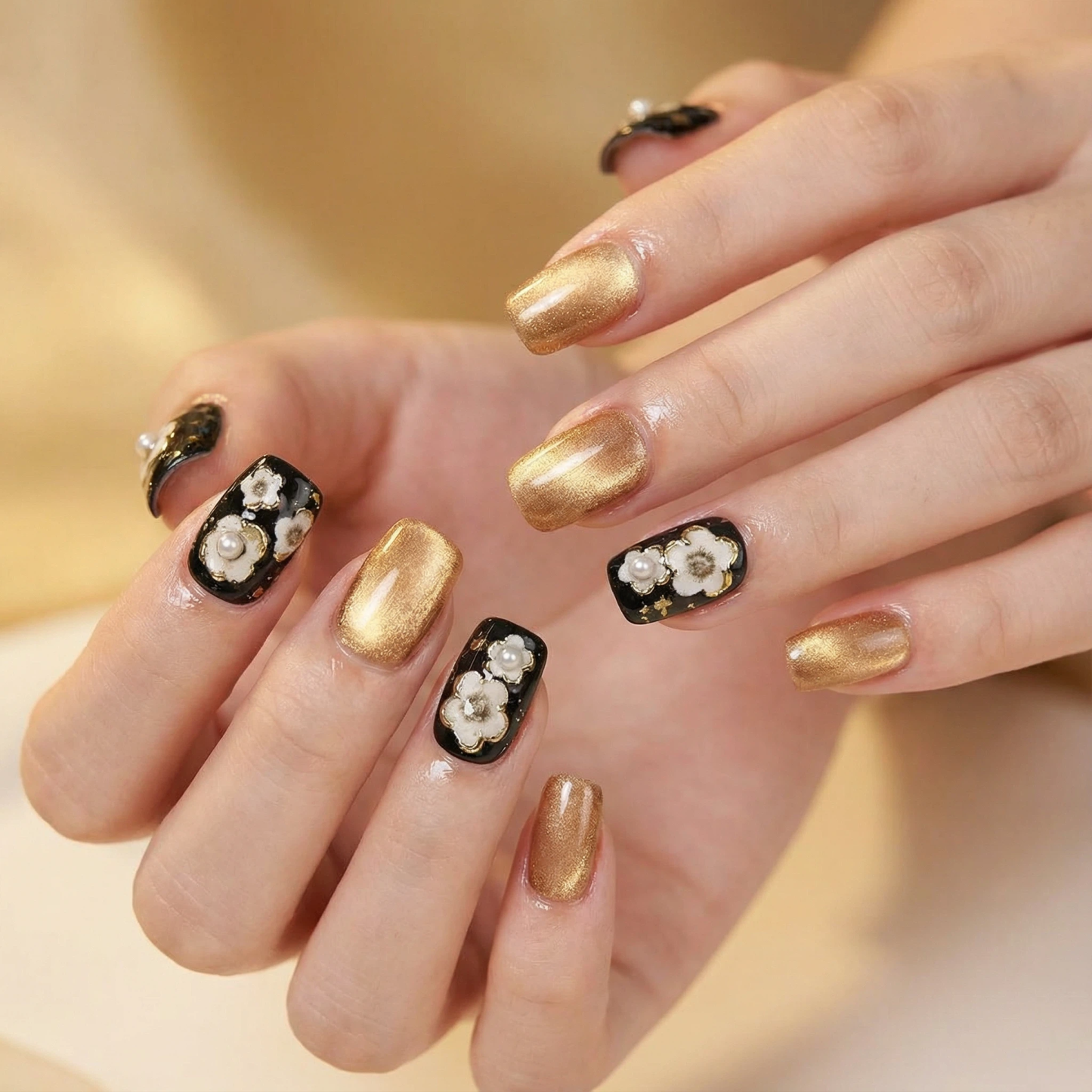 Black and gold press-on nails with white floral design and pearl accents on short square nails