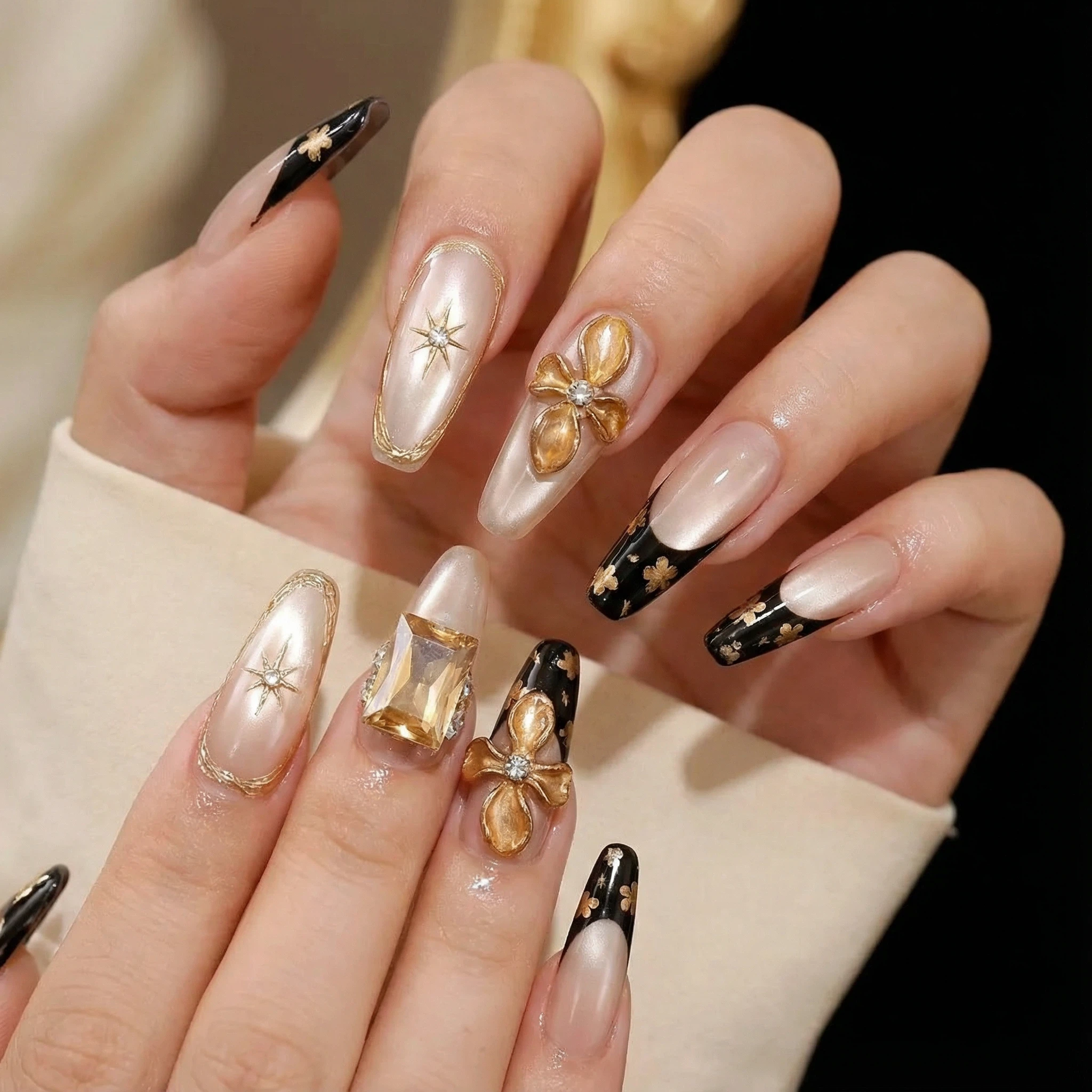 Hands wearing nude and black French tip press on nails with gold flower and gemstone accents.