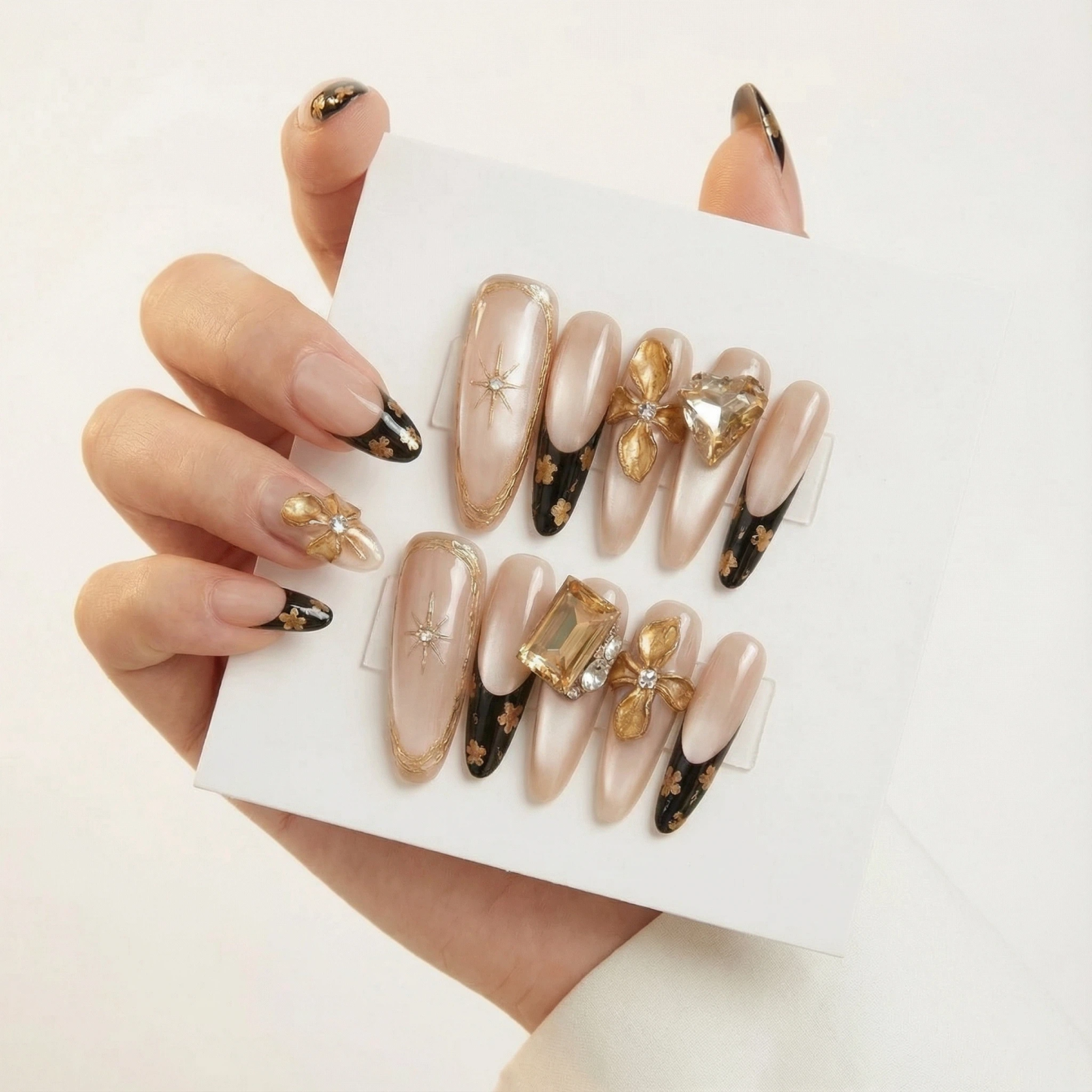 Nude and black French tip press on nails with gold 3D flower charms displayed on card.