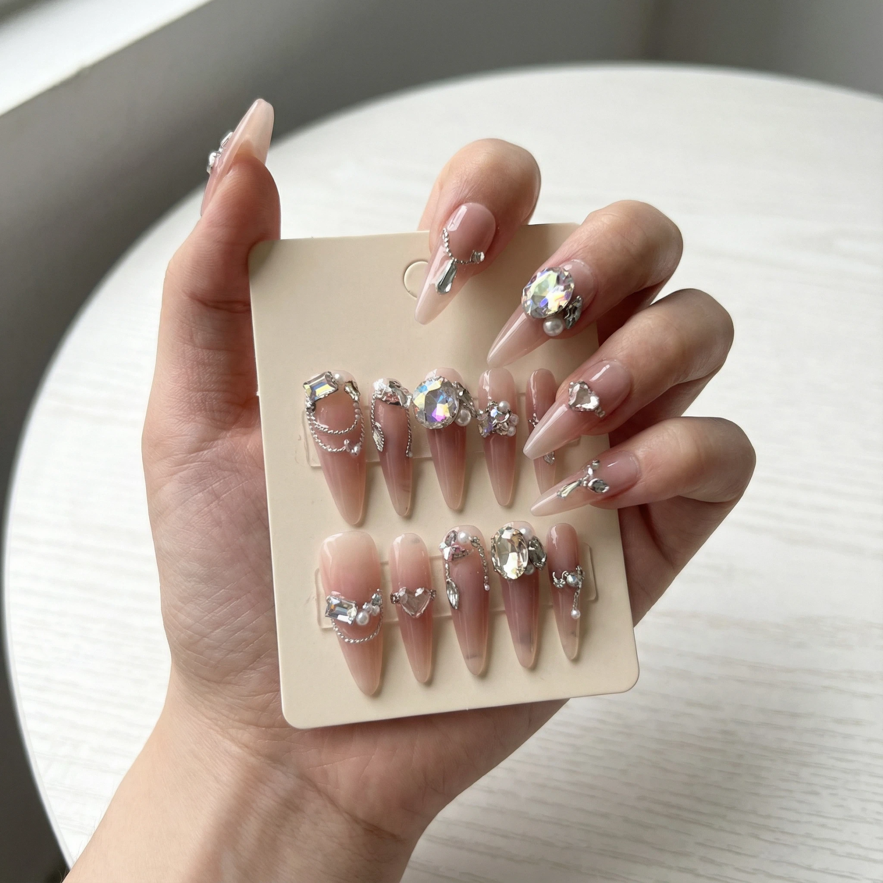 Hand holding a display card with a full set of nude pink long stiletto press on nails decorated with crystals and pearls.