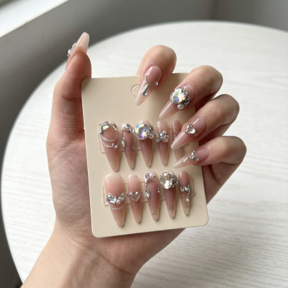 Hand holding a display card with a full set of nude pink long stiletto press on nails decorated with crystals and pearls.