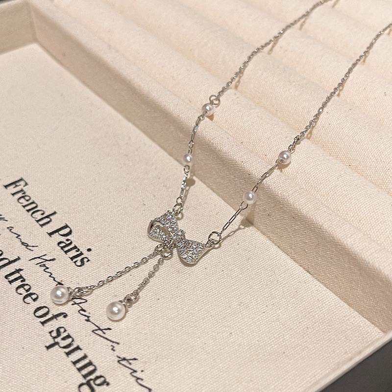 Silver-tone zircon bow necklace with pearl accents and dangling tassels displayed on light fabric