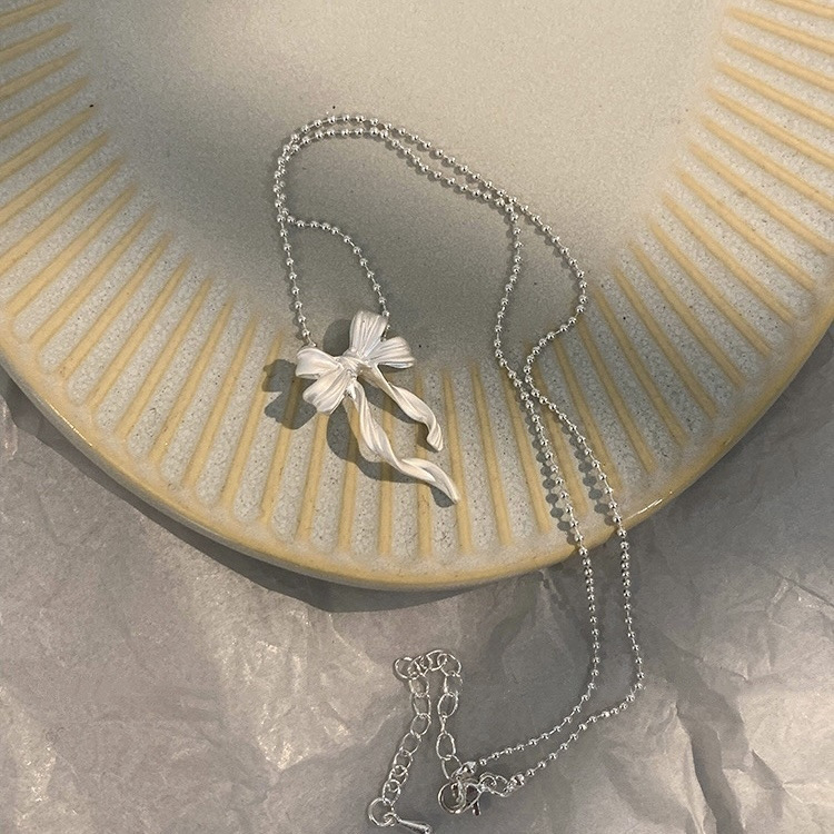 Silver ribbon bow pendant necklace arranged on a decorative ceramic plate with its chain and adjustable clasp visible