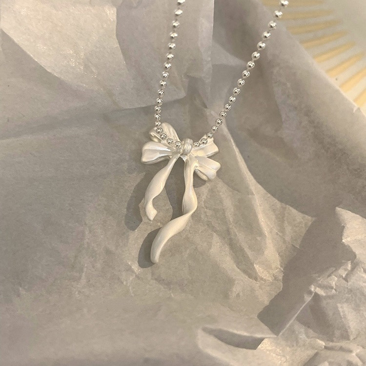 Silver ribbon bow pendant necklace displayed on a soft gray background, highlighting the textured bow and flowing tails