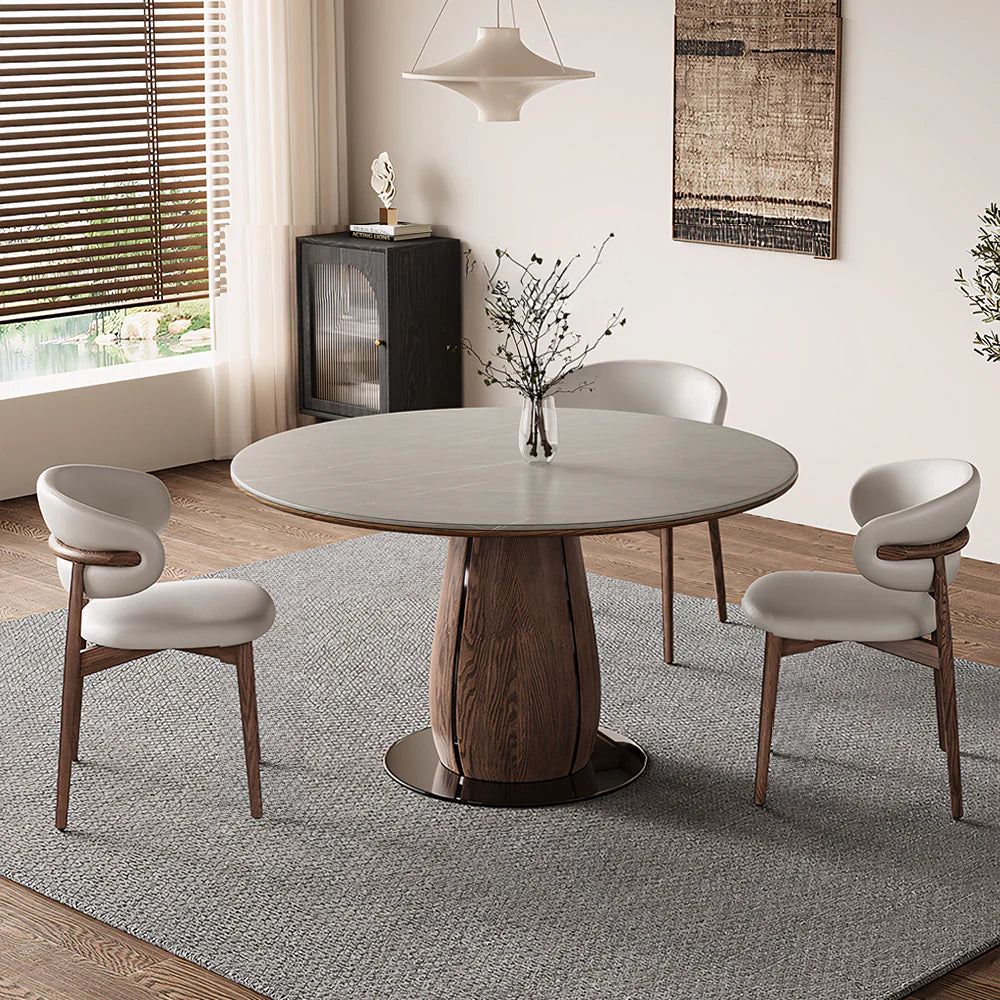 Round sintered stone dining table with a warm wood pedestal base, styled in a modern dining room with upholstered chairs and soft natural light.