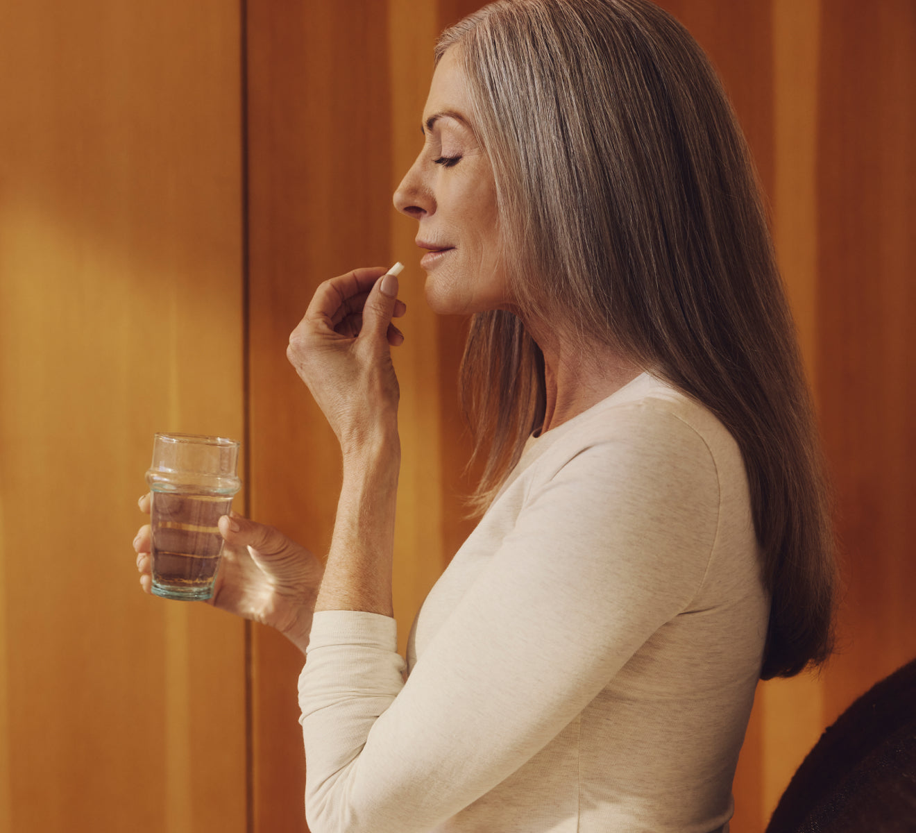 A woman holding a glass of water in one hand, and bringing a Tru Niagen supplement up to her mouth with the other.