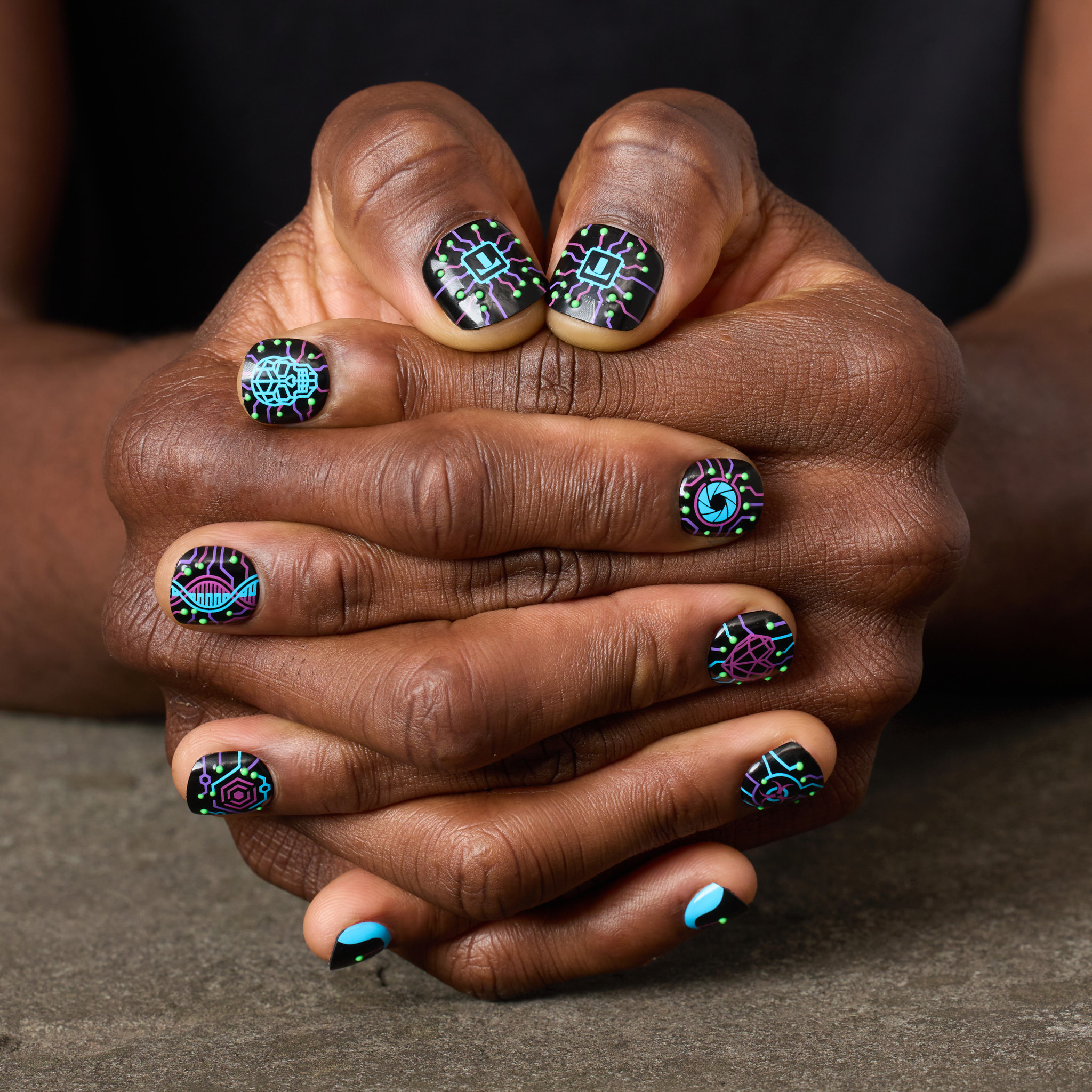 image of man's hands clasped, featuring the cyber neon press on nail set, which features a glowing circuitry crawling across a matte black base like code breaking through the system. Electric purple and cyan pathways power skull, heart, and eye icons that flicker like glitches in the simulation. 