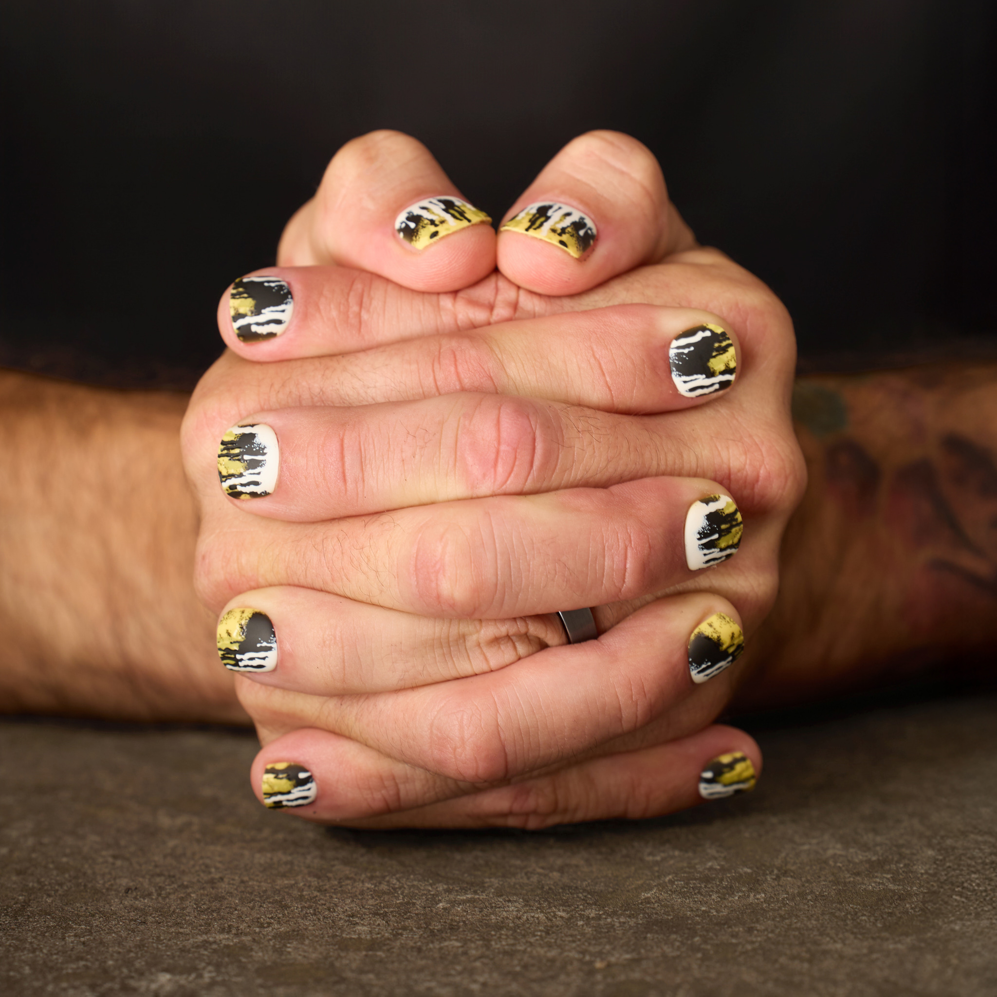 image of man's hands clasped featuring the toxic waste press on nail set. this set has a dripped and splattered design in a collision of neon yellow, jet black, and white.
