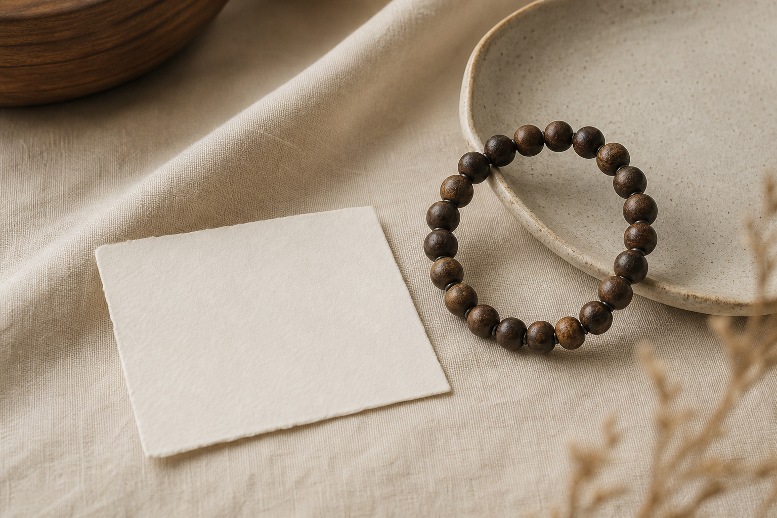 A dark wooden bead bracelet beside a blank card and ceramic dish on warm linen.