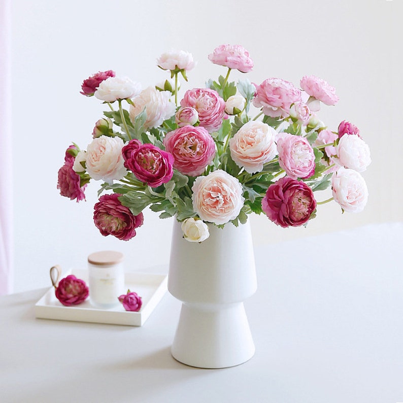 May include: A white ceramic vase with a wide base and a narrow neck, filled with a bouquet of pink and white artificial flowers. The flowers are arranged in a loose, natural style, and the vase is sitting on a white table.