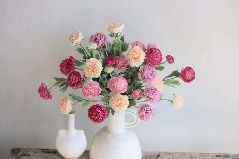 May include: A bouquet of pink, peach, and white ranunculus flowers in a white ceramic vase with a handle. The vase is sitting on a wooden surface.