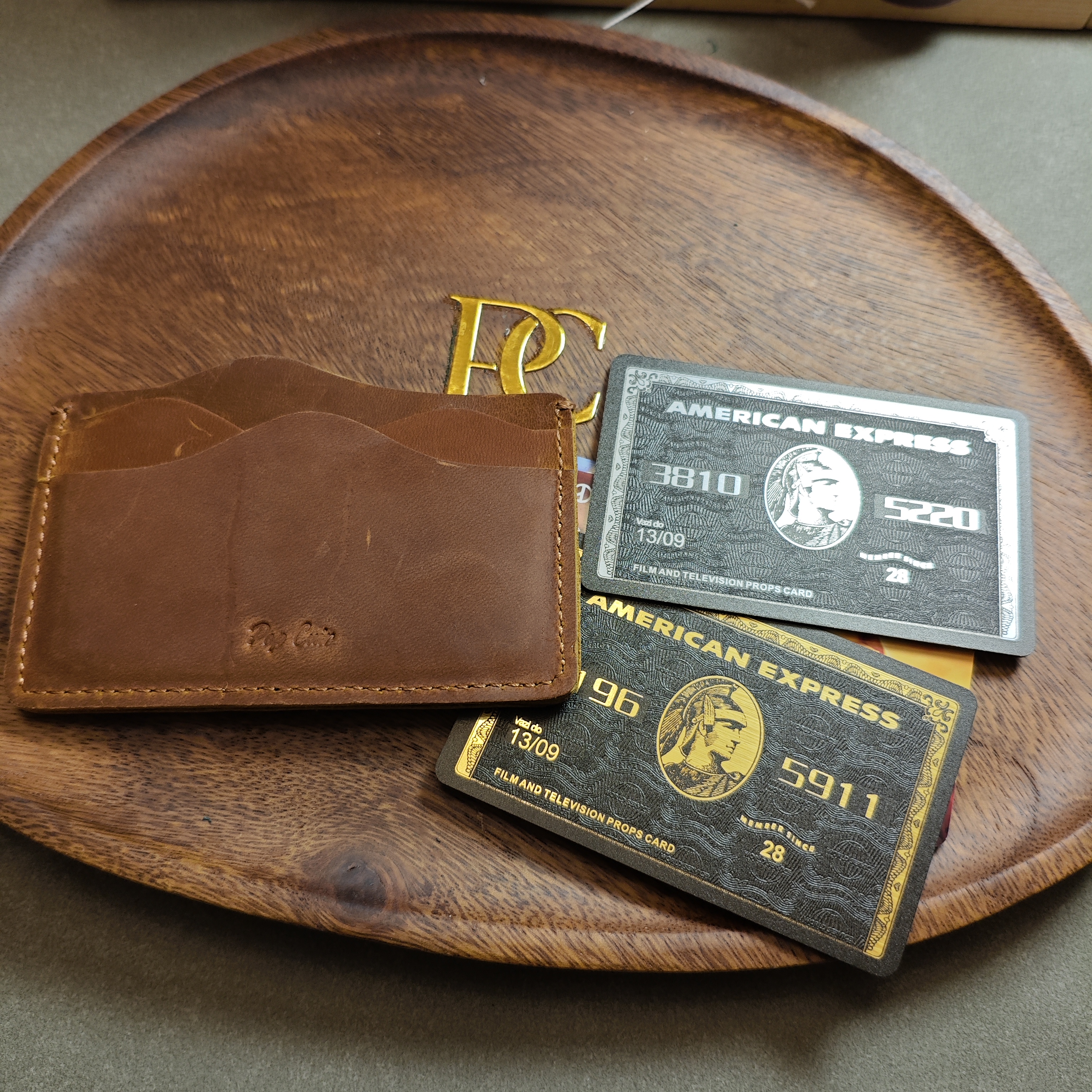 Open crazy horse leather card holder showing staggered 5-slot design with two American Express cards inserted in front pockets, displayed on wooden tray with Pop Cuir brass logo